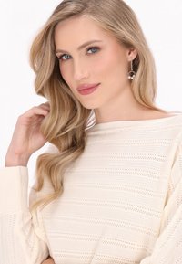 Jasno beżowy sweter dzianinowy w poziome paski z ściągaczami; modelka z długimi falującymi włosami i wiszącymi srebrnymi kolczykami z perełkami.
