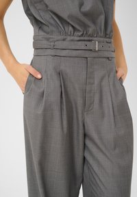 Pantaloni grigi su misura con fronte plissettato, gambe larghe e vita con cintura dotata di fibbia argentata e numerosi passanti.