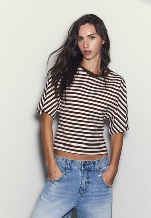 Femme aux longs cheveux foncés portant un t-shirt à manches courtes rayé marron et blanc et un jean bleu clair, les mains dans les poches.