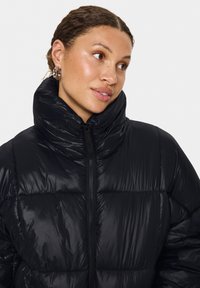 Svart puffjacka med hög, vadderad krage, glänsande yta, texturerat tyg och fullständig framdragkedja.