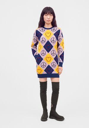 Femme portant une longue robe pull avec des motifs losanges jaunes, roses et bleus comportant des cœurs rouges et des signes de paix noirs, associée à des bottes noires montantes.