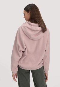 Vrouw met steil bruin haar in een lichtroze fleece jack met capuchon en donkergroene broek, met de rug naar de camera gekeerd tegen een effen achtergrond.