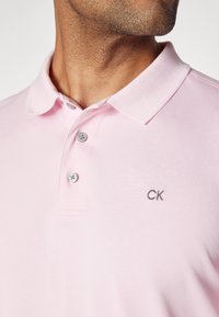 Ljusrosa polo skjorta i texturerad tyg, med klassisk krage, tre knappstängning och broderad "CK" logotyp på bröstet.