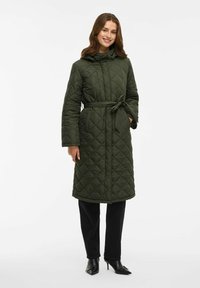 Femeie zâmbitoare îmbrăcată cu o haină lungă matlasată, de culoare verde măslinie, cu o curea, pantaloni negri și botine negre cu toc ascuțit, stand pe un fundal alb.