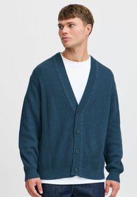 Mørk teal ribbestrikket cardigan med V-hals, lange ermer og fem-knaps frontlukking. Brukt over en hvit skjorte, vist mot en nøytral bakgrunn.
