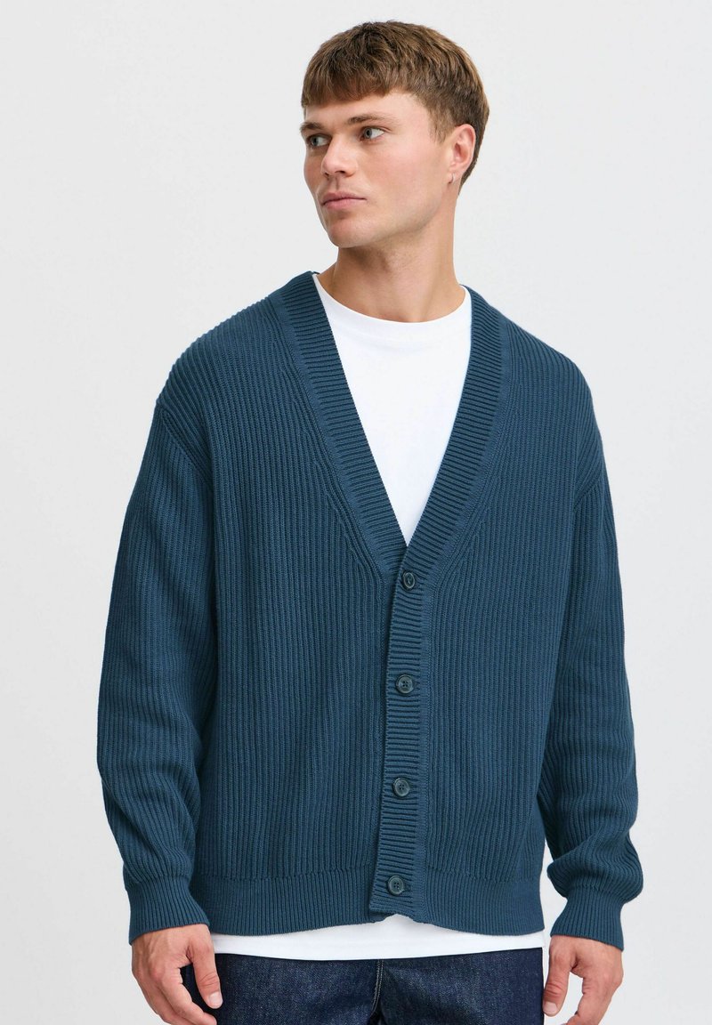 Mørk teal ribbestrikket cardigan med V-hals, lange ermer og fem-knaps frontlukking. Brukt over en hvit skjorte, vist mot en nøytral bakgrunn.