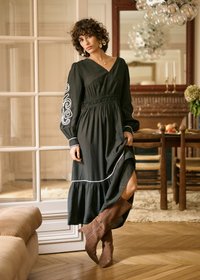 Dlouhé černé šaty s vyšívanými spirálovými vzory na rukávech, se staženým pasem a vrstveným lemem. Kombinované s hnědými semišovými kozačkami ke kolenům.