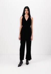 Svart sammetsjumpersuit med djup V-ringning, figurnära bodice, vida ben och knytning i midjan, kombinerad med spetsiga svarta ankelskor.