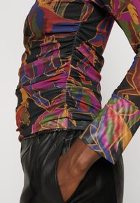 Langärmliges Oberteil in Schwarz mit buntem, abstraktem Blumenmuster. Strukturierter Stoff mit gerafften Seitennähten, kombiniert mit schwarzen Lederhosen.