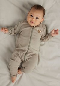 Bébé allongé sur un tissu gris clair, portant une grenouillère beige à fermeture éclair avec une petite illustration de renard sur la poitrine, regardant légèrement vers le haut.