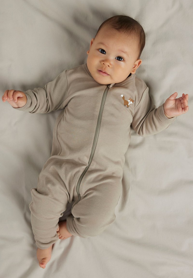 Bébé allongé sur un tissu gris clair, portant une grenouillère beige à fermeture éclair avec une petite illustration de renard sur la poitrine, regardant légèrement vers le haut.