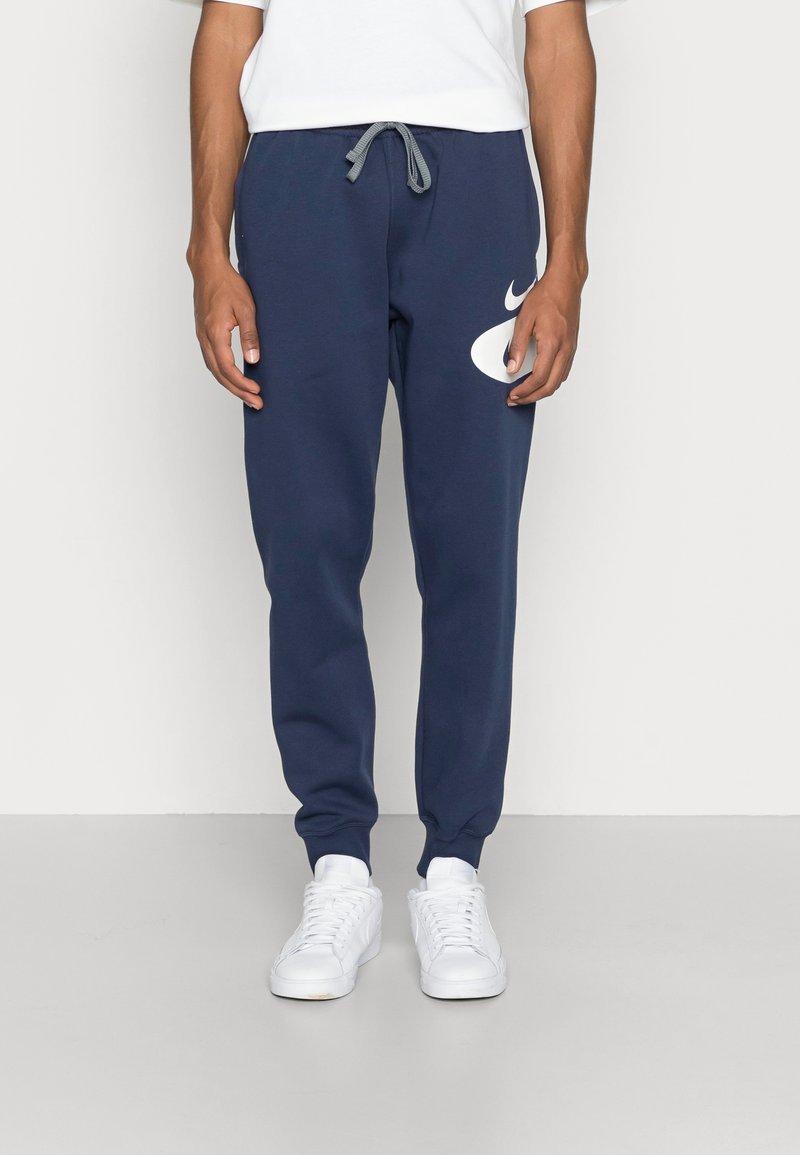 Uomo che indossa pantaloni jogger blu navy con il logo Nike bianco e sneakers bianche, in piedi su uno sfondo chiaro e uniforme.