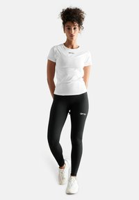 Witte getailleerde t-shirt met een geribbelde textuur en zwart logo, gecombineerd met hoge zwarte leggings met een logo. Schoeisel: lichtgekleurde sneakers.