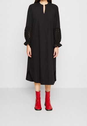 Mujer con un vestido midi negro con mangas abullonadas texturizadas y botas rojas hasta el tobillo, de pie frente a un fondo liso.