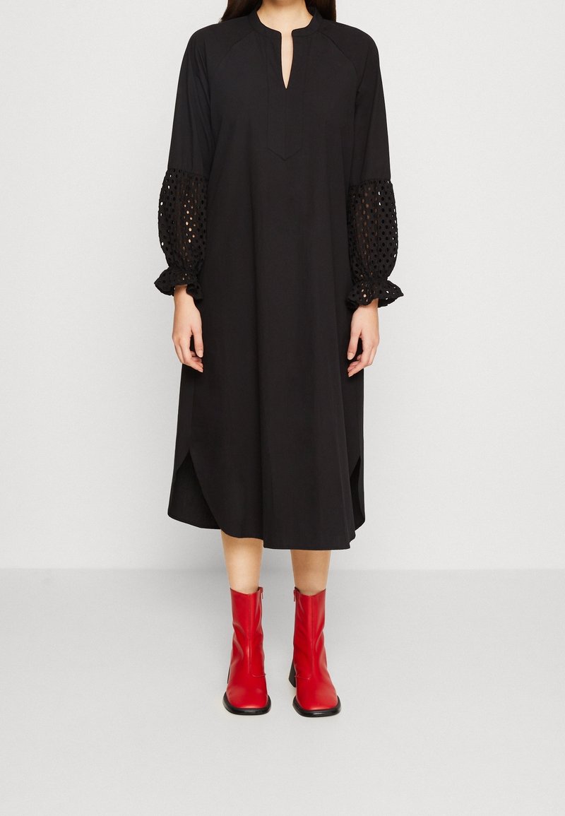 Femme portant une robe midi noire avec des manches ballon texturées et des bottines rouges, debout devant un fond uni.