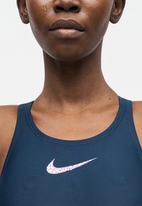 Κοντινό πλάνο ενός ατόμου που φοράει σπορ μπλούζα Nike σε ναυτικό μπλε με ροζ σχέδιο λογότυπου Nike Swoosh στο στήθος.