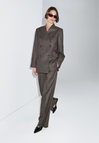 Completo marrone doppiopetto con pantaloni lunghi, caratterizzato da spalle strutturate e bottoni metallici. Abbinato a ballerine nere a punta e occhiali da sole.