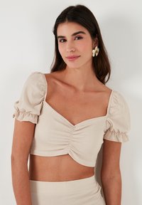 Gaiši beige kraupots tops ar pūkainajām piedurknēm, teksturētu audumu un priekšējo rušējuma detaļu. Apvienots ar saderīgu pieguļošu svārku.