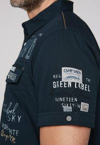 Navy short-sleeve shirt featuring various colorful embroidered patches and tags, with a button detail on the sleeve and contrasting stitching.