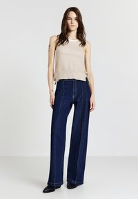 Giovane donna in piedi che indossa un top senza maniche beige in maglia, jeans a gamba larga blu scuro con cuciture frontali e scarpe a punta nere su uno sfondo neutro.