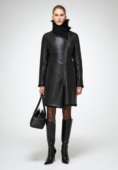 Donna che indossa un cappotto di pelle nera con colletto alto, stivali neri fino al ginocchio, mentre tiene in mano una borsa nera, in piedi davanti a uno sfondo semplice.