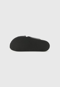 Sorte slip-on sandal med en teksturert gummisåle som har dype riller for grep og et glatt bånddesign på toppen.