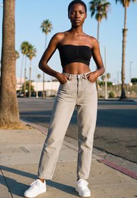 Fiatal nő fekete pánt nélküli felsőben és világosszürke farmerben áll a napsütötte, pálmafákkal szegélyezett utcán, kezét zsebében tartva.