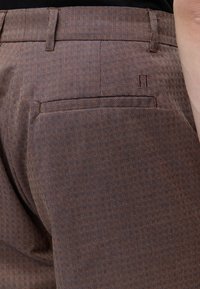 Gros plan sur un pantalon habillé marron à motifs montrant la poche arrière et les passants de ceinture avec un petit logo brodé au-dessus de la poche.