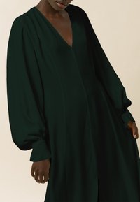 Largo vestido verde oscuro con un profundo escote en V, mangas voluminosas y una silueta fluida. Tela suave con un brillo sutil.
