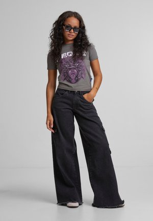 Szary t-shirt z grafiką w purpurowym kolorze, w połączeniu z szerokimi czarnymi dżinsami i ciemnymi okularami przeciwsłonecznymi, stojący na neutralnym tle.