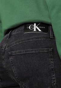 Jeans de mezclilla negra con costuras visibles, con una etiqueta de Calvin Klein en la pretina y una sudadera verde que los cubre parcialmente.
