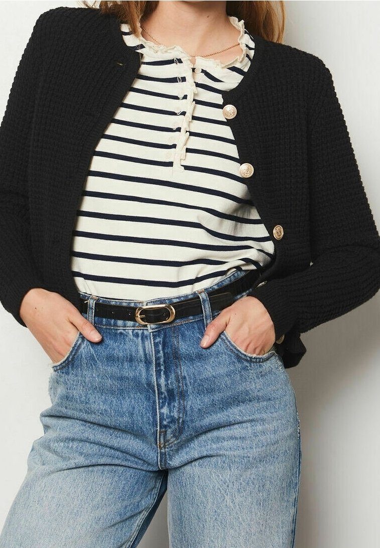 Schwarze Strickjacke mit goldenen Knöpfen, über einem gestreiften Hemd getragen. Hoch taillierte blaue Jeans und ein schwarzer Gürtel vervollständigen das Outfit.