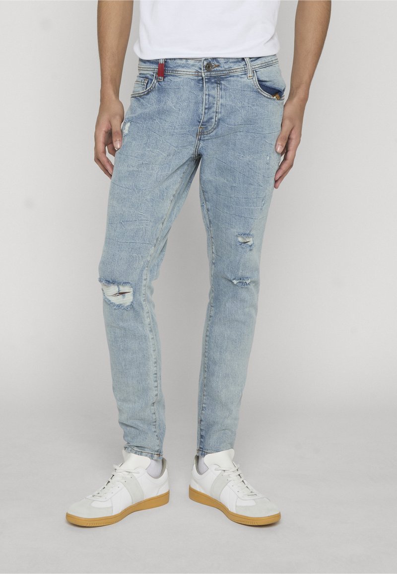 Muž v svetlom modrom opotrebovanom rifľovom a bielych teniskách stojaci na jednofarebnom pozadí.