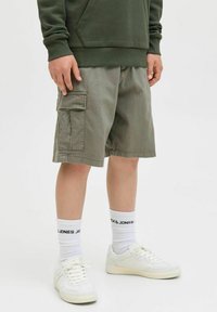 Khaki cargo kratke hlače z bočnimi žepi, v kombinaciji z belimi športnimi copati in soknami do gležnjev z črnim logotipom. Tkanina se zdi lahka.