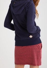 Sweat à capuche bleu marine avec des pois blancs et roses, doté d'une capuche et d'une fin en côtes. Associé à une jupe rouge chinée, présentant un design superposé.