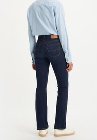 Jean en denim bleu foncé avec des jambes évasées, taille haute et coutures dorées contrastantes. L'arrière présente une étiquette logo rouge et des poches courbées.