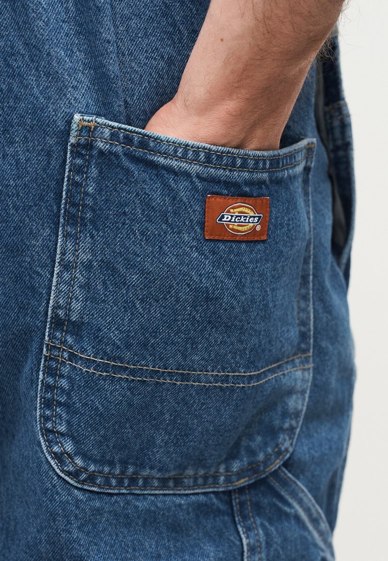 Mano inserita nella tasca posteriore di pantaloni di jeans blu con visible patch del logo del marchio Dickies.