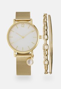 Montre à bracelet en maille dorée avec un cadran blanc, ornée d'index en or et d'un petit charme avec une pierre transparente. Comprend deux bracelets dorés.