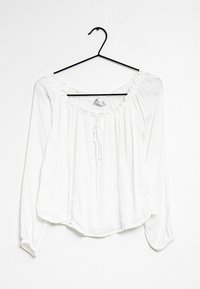 Blusa bianca a maniche lunghe con scollatura rotonda arricciata, dettaglio con fiocco, tessuto texturizzato e pieghe verticali lungo la parte anteriore.