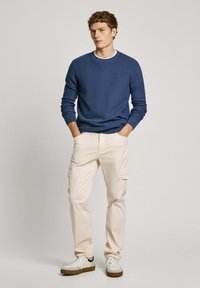 Maglione blu lavorato a maglia con scollo rotondo, pantaloni cargo crema e sneakers bianche con suole marroni, che mostrano uno stile casual.