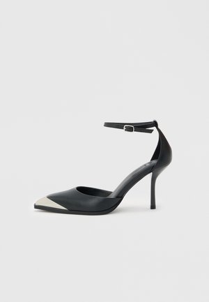 Zwarte hoge hak met puntige neus, zilverkleurige metalen neuskap en enkelbandje met gespsluiting op een witte achtergrond.