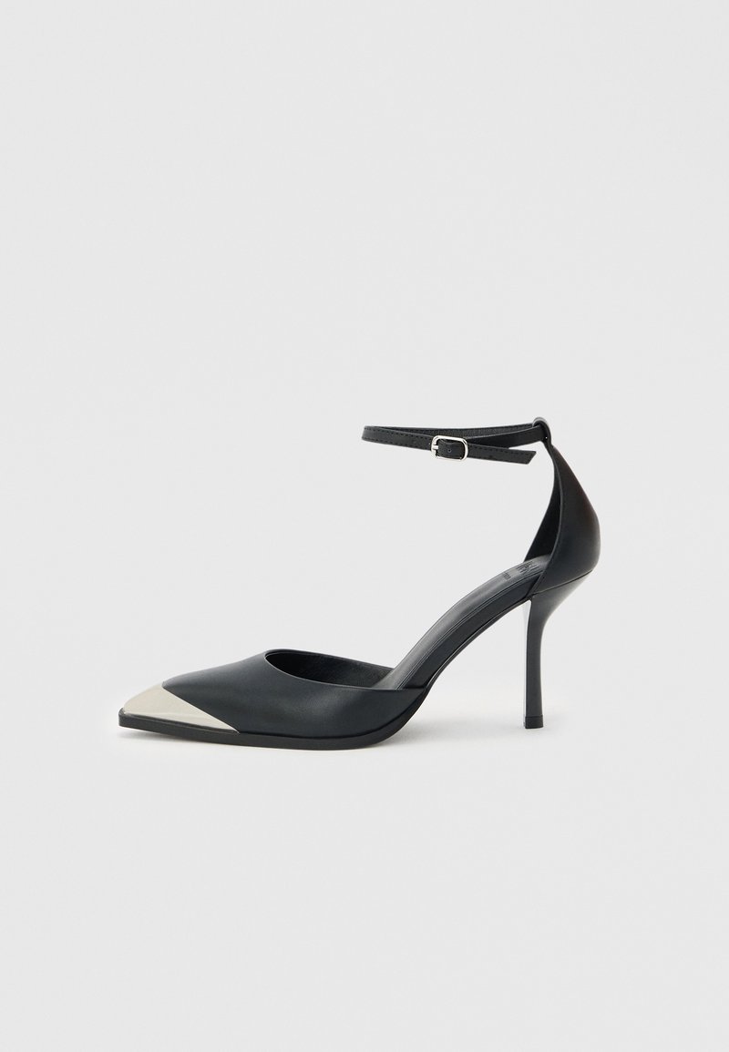 Μαύρο γυναικείο παπούτσι με ψηλό τακούνι και μυτερή μύτη, με ασημί μεταλλική επένδυση στη μύτη και λουράκι στον αστράγαλο με κούμπωμα, σε λευκό φόντο.