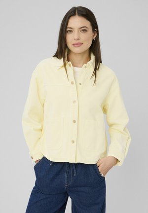 Jeune femme aux longs cheveux foncés portant une veste pale jaune boutonnée et un pantalon en denim bleu foncé, mains dans les poches, fond neutre.