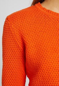 Pull en maille orange avec un motif texturé, col rond et tissu de poids moyen, offrant une coupe confortable et des poignets côtelés.
