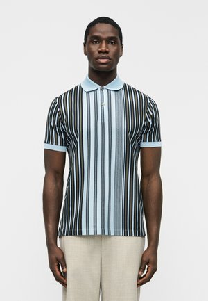 Mężczyzna ubrany w niebiesko-czarną koszulkę polo w pionowe paski z krótkim rękawem oraz jasnobeżowe spodnie, stojący na jednolitym tle.