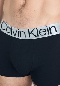 Zwarte katoenen boxershorts met een zachte, brede grijze tailleband met "Calvin Klein" in zwarte letters. Glad textuur en aansluitend ontwerp.
