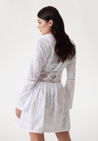 Weißes besticktes Kleid mit langen Glockenärmel, Spitzenakzenten an der Taille, floralen Mustern und gewelltem Saum. Leichtes und strukturiertes Material.