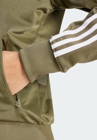 Svetle olivová bunda s ribbenými manžetami, bočním zipovým kapsou a třemi bílými proužky na levém rukávu. Hladká, textura látka.