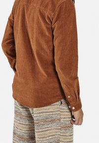 Camisa de pana marrón con mangas largas y puños con botones, combinada con pantalones beige y marrones con textura y estampado.