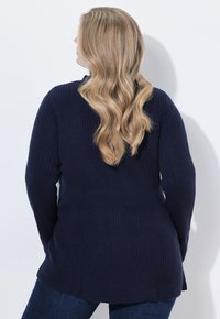 Kvinna med långt vågigt blont hår, iförd en marinblå stickad tröja och mörka jeans, vänd mot en vit vägg.